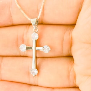 Op de afbeelding: Een zilverkleurige kruishanger aan een fijne schakelketting. Het kruis heeft een gepolijste metalen afwerking en is voorzien van vier ronde, heldere, gefacetteerde stenen aan de bovenkant, onderkant en de uiteinden van de zijarmen.