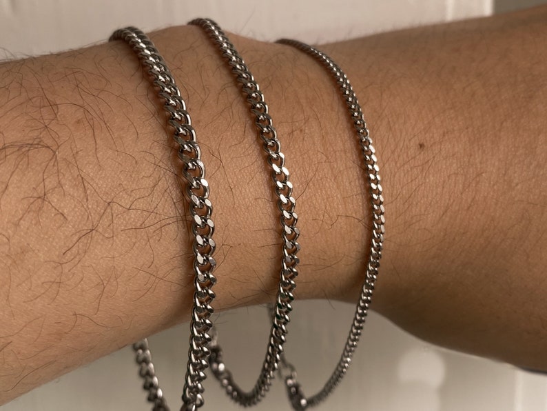 May include: Three silver chain bracelets of different widths on a person's wrist.