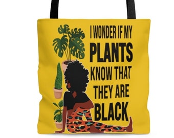 May include: A yellow tote bag with a black handle and a black and white illustration of a woman with plants. The text on the bag reads "I wonder if my plants know that they are black."