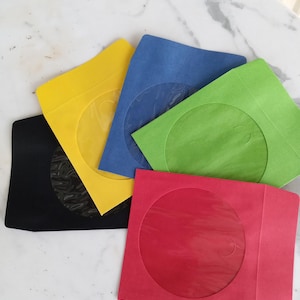 May include: A collection of colorful CD/DVD sleeves in yellow, blue, green, red, and black. Each sleeve has a circular transparent window. The sleeves are arranged on a white marble-like surface.