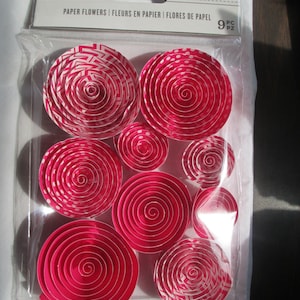 May include: Nine pink paper flowers with white patterns. The flowers are in a clear plastic package with a white label that says "Darice Paper Flowers | Fleurs en Papier | Flores de Papel 9 pc pz".