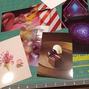 May include: A collection of various postcards and prints, including images of a flamingo pool float, a woman in a yellow swimsuit, a geometric design, a Pi symbol, and chocolates. The items are arranged on a green cutting mat.