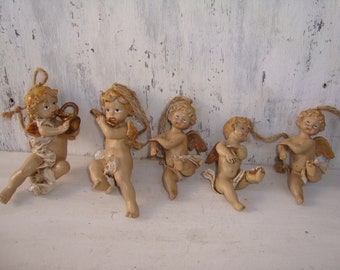 Puede incluir: Cinco pequeñas figuritas de ángeles beige con alas y lazos de cuerda para colgar. Los ángeles están en varias poses, algunos sosteniendo instrumentos musicales.