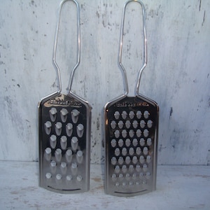 May include: Two silver metal cheese graters with handles. The grater on the left has a coarse grate and the grater on the right has a fine grate. Both graters have "STAINLESS STEEL" printed on them.
