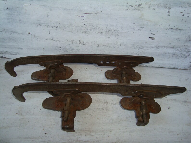 May include: A pair of vintage rusty metal ice skate blades. The blades are attached to a metal frame with a screw and a metal plate. The blades are in good condition, but show signs of wear and tear.