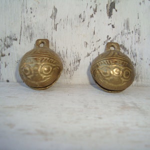 May include: Two small, gold-colored bells with intricate designs. The bells have a rounded shape and a small loop at the top for hanging.
