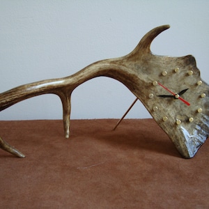 May include: A unique clock made from a deer antler with a brown and white antler base and a clock face with gold bullet casings for numbers. The clock hands are black and red.