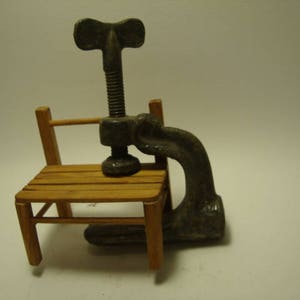 May include: A vintage metal clamp with a wooden handle, attached to a small wooden chair. The clamp has a screw mechanism and a metal handle.