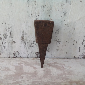 May include: A rusty metal anvil with a square top and a pointed bottom. The anvil is sitting on a white wooden surface.