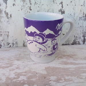 Könnte beinhalten: Lilafarbene Keramiktasse mit einem weißen Schneemann und einem Skifahrer-Motiv. Die Tasse hat den Schriftzug "Milka" auf der Seite.