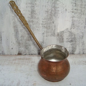 May include: A copper and brass ladle with a long, ornate handle. The ladle has a round, bulbous body and a wide, shallow bowl. The handle is decorated with a repeating pattern of loops and swirls.