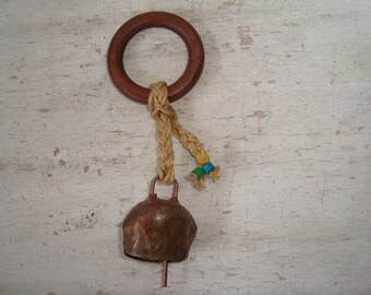 May include: A rustic bell with a copper-colored body and a wooden ring at the top. The bell is suspended by a natural rope, adorned with small blue and yellow beads. The background is a distressed white surface.