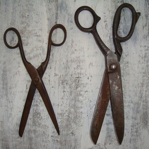 May include: Two pairs of vintage scissors with rusted metal blades. The larger pair has a longer handle and is open. The smaller pair is closed and has a shorter handle.