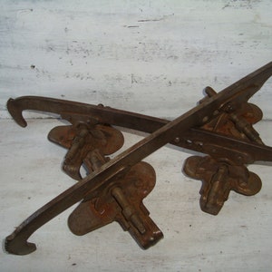 May include: A pair of vintage rusty metal ice skate blades with a hook on each end. The blades are attached to a metal base with screws.