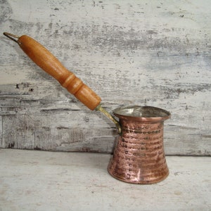 May include: A copper coffee pot with a hammered finish and a wooden handle. The pot has a small spout and a rounded base.