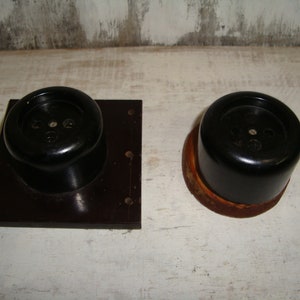 Puede incluir: Dos tapas de toma de corriente de baquelita negra con tres agujeros cada una. La tapa de la izquierda está montada en una placa cuadrada negra. La tapa de la derecha está sobre una superficie marrón.