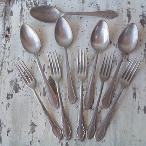 May include: A set of vintage silverware, including spoons and forks, arranged in a fan shape on a white wooden surface. The silverware is made of silver and has a tarnished finish.