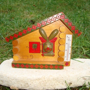 May include: A wooden thermometer shaped like a house with a rabbit peeking out of a window. The thermometer has a red and green diamond pattern on the roof and a red and green border around the base. The thermometer is on a white rock.