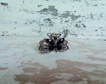 May include: A silver butterfly hair clip with black rhinestones. The clip is on a white wooden background.