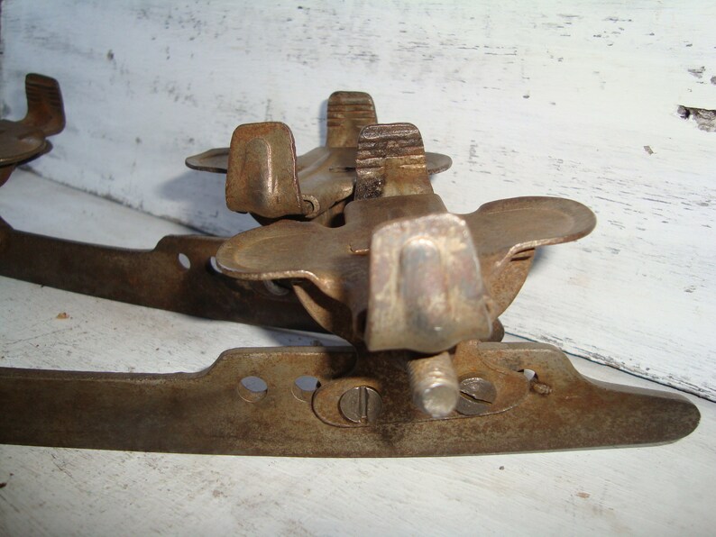 May include: A pair of vintage rusty metal ice skate blades with a metal toe stop. The blades are attached to a metal frame with a screw.