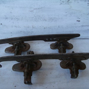 May include: A pair of vintage metal ice skate blades with a rusty finish. The blades are attached to a metal frame with a screw and a lever.