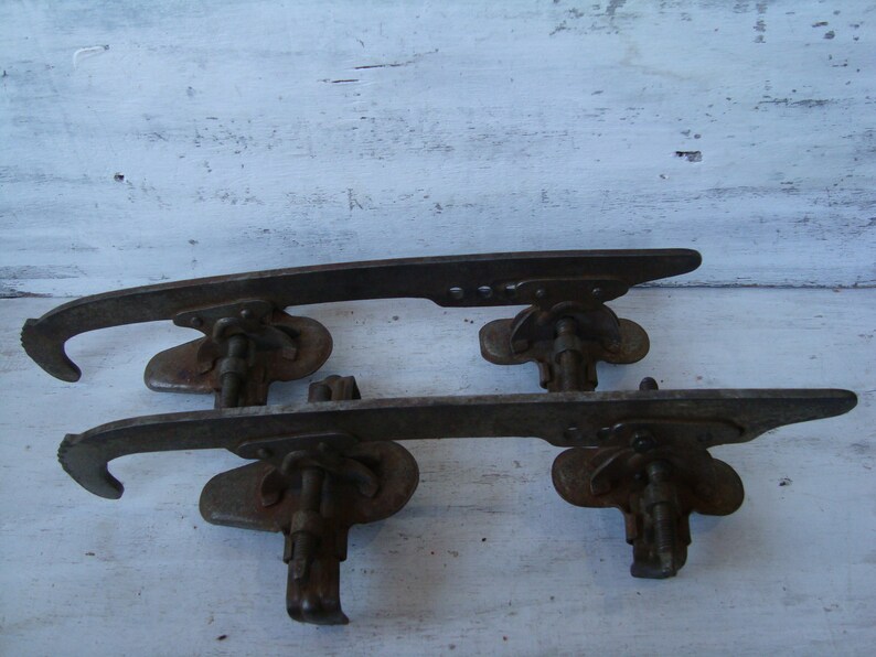 May include: A pair of vintage metal ice skate blades with a rusty finish. The blades are attached to a metal frame with a screw and a lever.