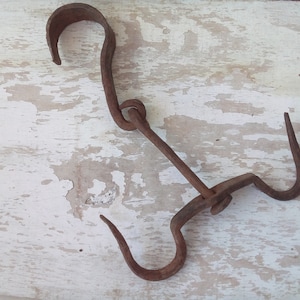 May include: A rusty metal hook with two loops. The hook is made of a single piece of metal that is bent into a U-shape with a loop at the top and a loop at the bottom. The hook is on a white wooden surface.