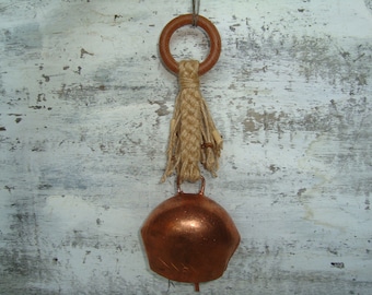 May include: A copper cowbell with a brown wooden ring and a natural fiber tassel hanging from a whitewashed wooden surface.