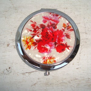 May include: A round silver compact mirror with a floral design. The design features red, pink, and yellow flowers on a white background.