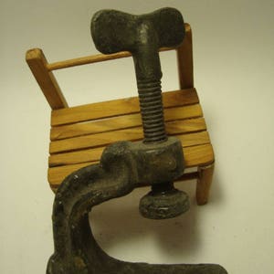 May include: A vintage metal clamp with a T-shaped handle. The clamp is made of dark grey metal and has a smooth, worn surface. It is resting on a wooden surface.