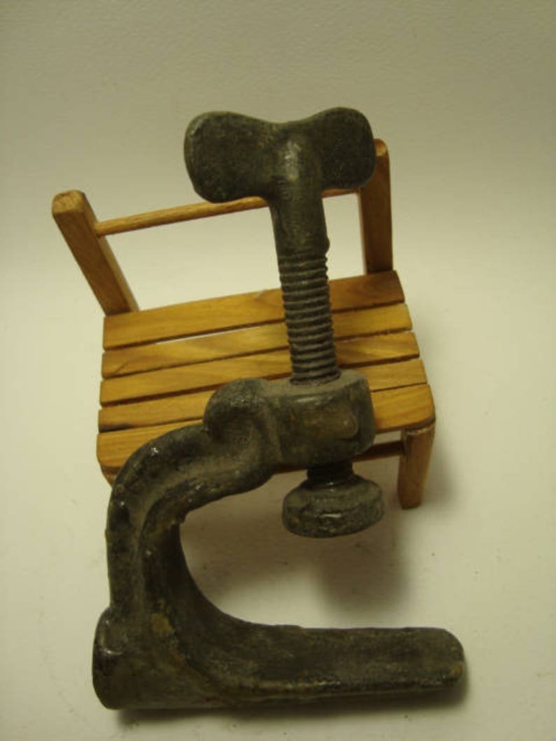 May include: A vintage metal clamp with a T-shaped handle. The clamp is made of dark grey metal and has a smooth, worn surface. It is resting on a wooden surface.
