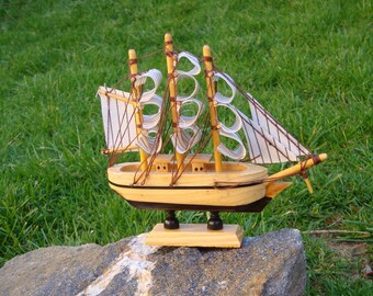 May include: A wooden model sailboat with tan sails and a light brown hull. The miniature ship has three masts and is mounted on a small wooden base. The model is displayed on a gray rock against a backdrop of green grass.