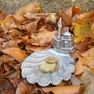 Peut inclure: Une figurine miniature en argent d'une mosquée avec une base décorative en forme de coquille, entourée de pièces d'or, reposant sur un lit de feuilles d'automne brunes.