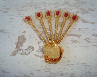 May include: Set of six gold-tone teaspoons with red gem-like accents. The spoons have ornate floral designs on the bowls.