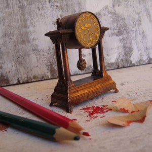May include: A small, antique-style, gold-coloured clock with a pendulum. The clock is sitting on a white surface with red and brown pencil shavings.