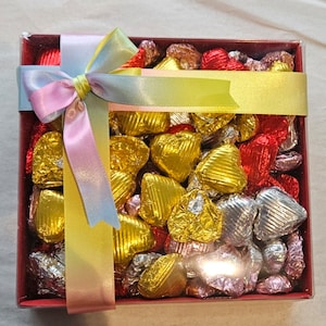 May include: A red box filled with heart-shaped chocolates wrapped in gold, silver, and pink foil. The box is tied with a multi-coloured ribbon.