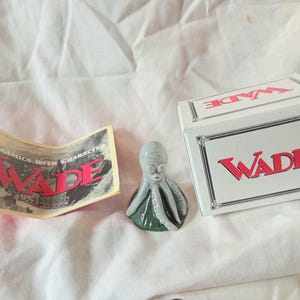 May include: A small ceramic octopus figurine with a light grey head and dark green tentacles, accompanied by a white rectangular box with the red text WADE and a paper insert featuring the black and white text WADE CERAMICS WITH CHARACTER.