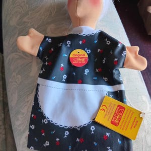 May include: A Steiff hand puppet of an elderly person with white hair and glasses, wearing a black dress with a white floral pattern and a white apron. The puppet features a round red Original Steiff tag and a yellow informational product tag.
