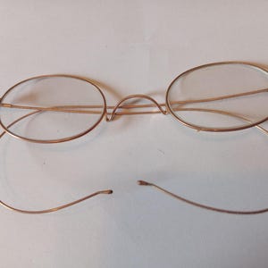 Könnte beinhalten: Antike goldfarbene Brille mit ovalen Gläsern. Die Drahtgestelle haben einen zarten, gebogenen Steg und Bügel. Die Bügel sind abgetrennt. Ein Vintage-Brillenstück.