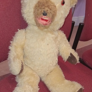 May include: A vintage teddy bear with cream-coloured fur, a brown snout, and red eyes. The bear is seated on a red chair. Its paws and feet are brown. A classic plush toy.