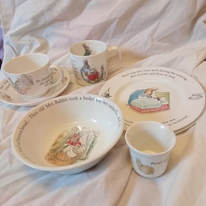 May include: A white ceramic Peter Rabbit children's dinnerware set including a bowl, a plate, two mugs with saucers, and an egg cup. Each piece features colourful illustrations of rabbits and printed text from the story, including phrases like Peter was not very well during the evening and Then old Mrs. Rabbit took a basket and her umbrella.