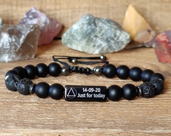 May include: A black beaded bracelet featuring matte onyx spheres, textured lava stone beads, and a central rectangular black metal charm. The charm displays a white triangle symbol, the date 14-09-20, and the text Just for today.
