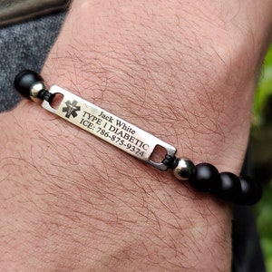 Op de afbeelding: Een medische alarmarmband met matzwarte kralen en een rechthoekige zilverkleurige metalen plaat. De plaat toont het symbool van de ster van het leven en de gegraveerde tekst: Jack White, TYPE 1 DIABETIC, ICE: 786-875-9374.