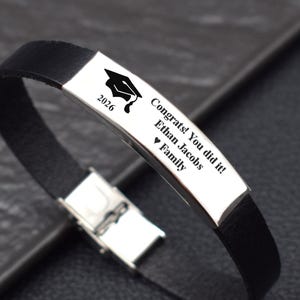 May include: Black leather bracelet with a silver plate engraved with a graduation cap, the year 2026, and the text "Congrats! You did it! Ethan Jacobs ❤ Family". The bracelet has a silver clasp.