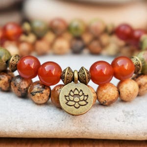 Op de afbeelding: Twee armbanden met bruine, groene en oranje kralen en een gouden bedel met een lotusbloemmotief.