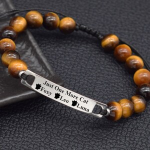 May include: A beaded bracelet with tiger eye stones and a silver bar. The bar is engraved with the text "Just One More Cat" and the names "Foxy, Leo, Luna" with paw prints. The bracelet is on a black surface.