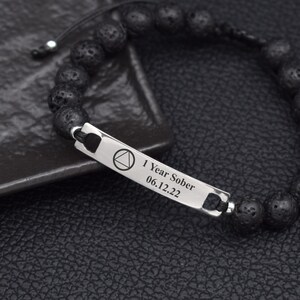 May include: Black lava stone bead bracelet with a silver-tone rectangular charm. The charm is engraved with the text "1 Year Sober" and the date "06.12.22", along with a triangle symbol. The bracelet is on a dark surface.