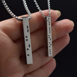 May include: Two silver-toned vertical bar pendants suspended from matching metal chains. Each rectangular bar features a series of black engraved dots and dashes arranged in a vertical pattern, resembling Morse code, with a polished metallic finish.