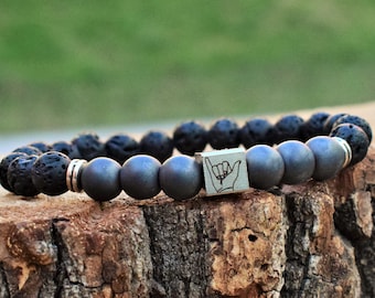 May include: A black lava rock and hematite beaded bracelet with a silver charm featuring a hand gesture.