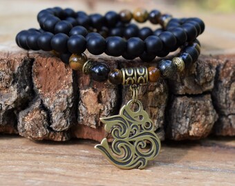 Könnte beinhalten: Ein Armband aus schwarzen Onyxperlen mit einem Akzent aus Tigerauge- und Hämatitperlen. Das Armband hat einen goldfarbenen Om-Anhänger.